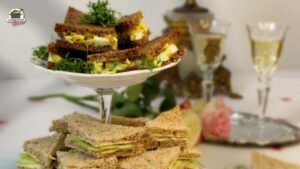 Etagere mit Ei-Kresse- und Gurken-Sandwiches. Im Hintergrund stehen zwei Likörgläser