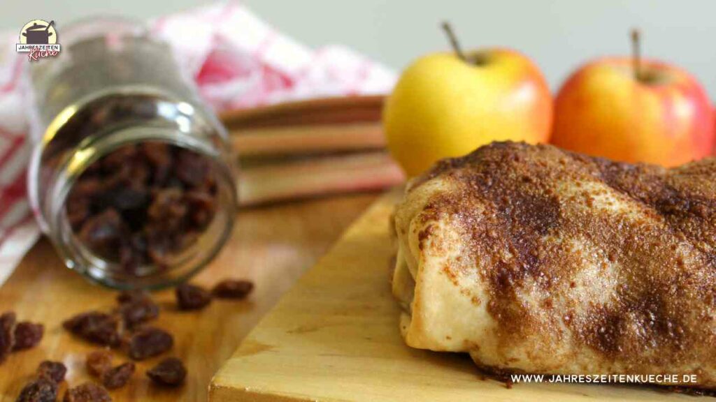 Ein Apfel-Rhabarber-Strudel mit Rosinen liegt einem Holzbrett. Im Hintergrund sind zwei Äpfel
