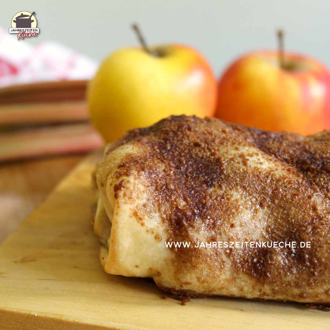Ein Apfel-Rhabarber-Strudel mit Rosinen liegt einem Holzbrett. Im Hintergrund sind zwei Äpfel
