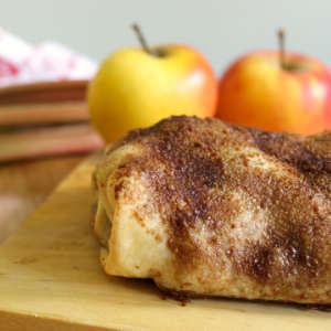 Ein Apfel-Rhabarber-Strudel mit Rosinen liegt einem Holzbrett. Im Hintergrund sind zwei Äpfel