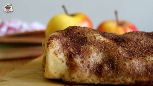 Vor zwei Äpfeln liegt ein Apfel-Rhabarber-Strudel mit Rosinen auf einem Holzbrett.