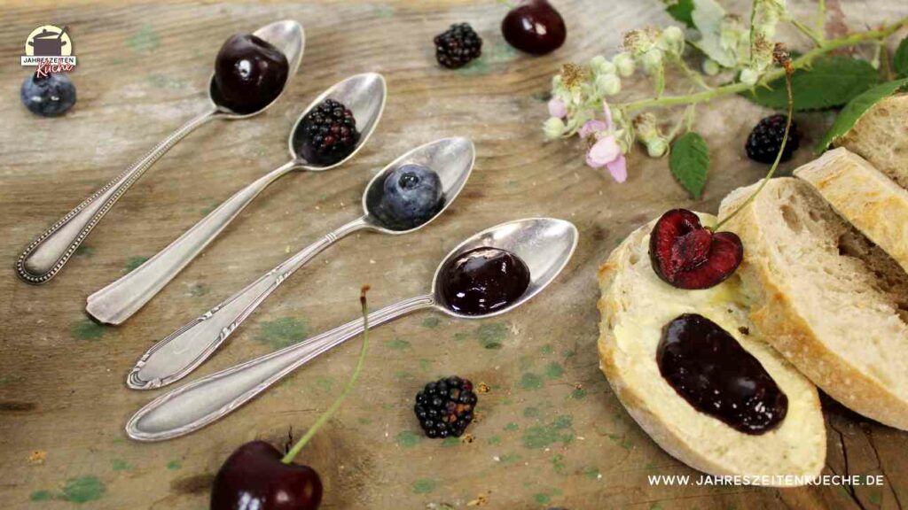 Neben silbernen Teelöffeln, auf denen ein Klacks Brombeer-Heidelbeer-Kirsch-Marmelade mit Rosenblüten ist liegt eine Scheibe Weißbrot mit einer halbierten Kirsche.