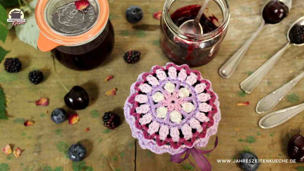 3 Gläser mit Brombeer-Heidelbeer-Kirsch-Marmelade stehe auf einem groben Holztisch. Ein Glas ist offen, eins ist mit einem Glasdeckel verschlossen und das dritte Glas ist mit einem gehäkelten Mandala abgedeckt.