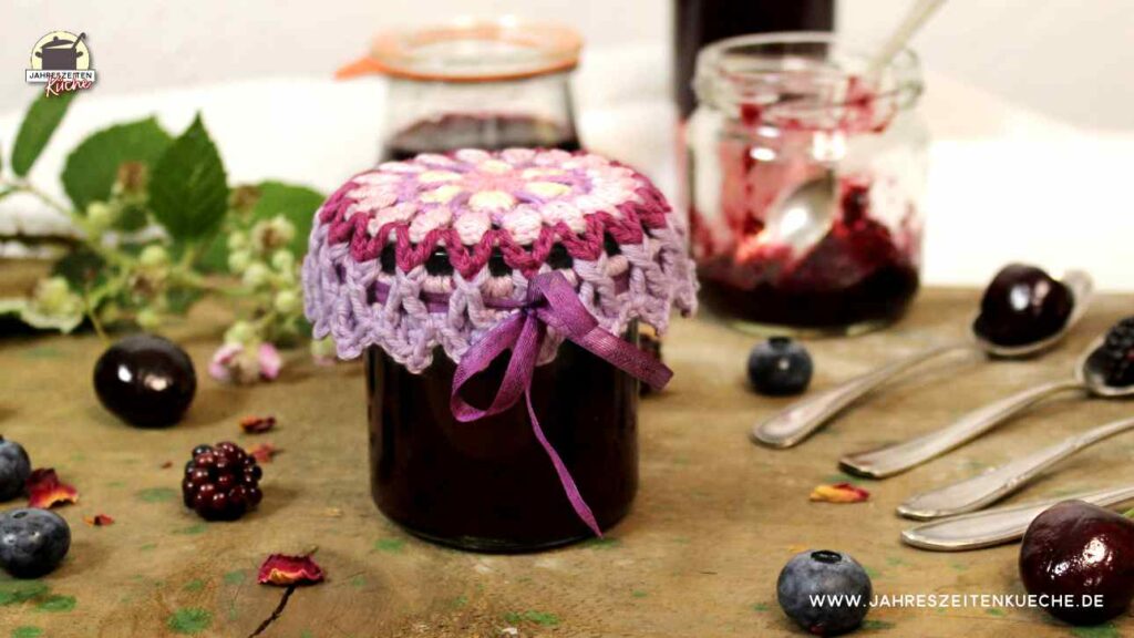 Auf einem groben Holztisch steht ein Glas Brombeer-Heidelberr-Kirsch-Marmelade. Im Hintergrund stehen zwei weitere Gläser.
