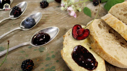 Neben silbernen Teelöffeln, auf denen ein Klacks Brombeer-Heidelbeer-Kirsch-Marmelade mit Rosenblüten ist, liegt eine Scheibe Weißbrot mit einer halbierten Kirsche.