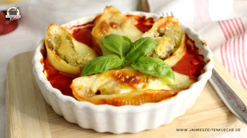 Auf einem Holzbrett stehe eine weiße Auflaufform mit gefüllter Muschelpasta im Tomatensugo