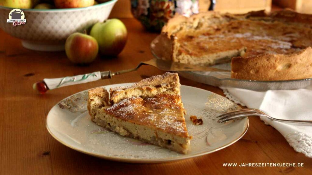 Auf einem Teller mit Goldmuster ist ein Stück Apfel-Käsekuchen. Im Hintergrund steht eine Schale mit Äpfeln