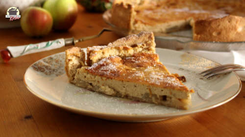 Auf einem Teller mit Goldmuster ist ein Stück Apfel-Käsekuchen, das mit Puderzucker bestäubt ist.