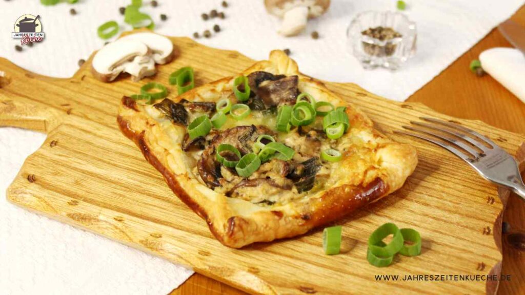 Ein Blätterteig-Snack mit Champignons und Frühlingszwiebeln liegt auf einem Holzbrett