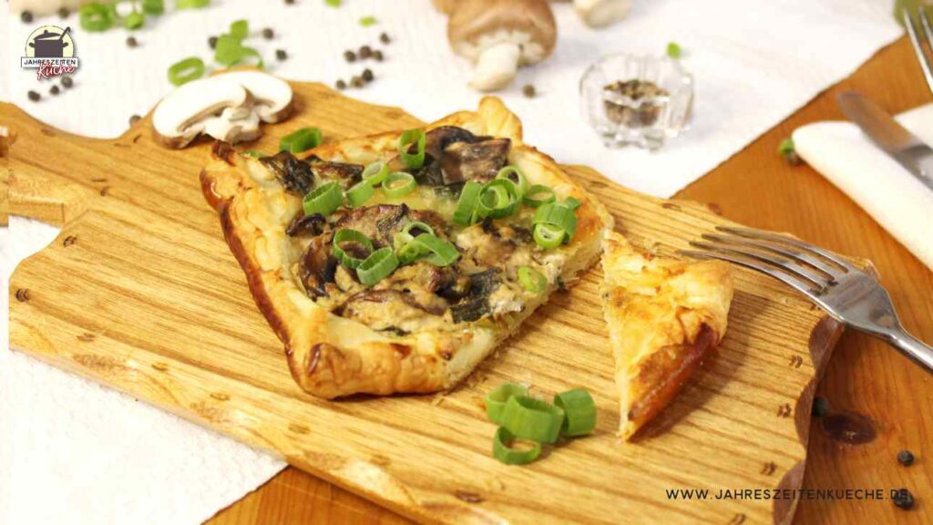 Auf einem Holzbrett liegt ein Blätterteig-Snack mit Champignons und Frühlingszwiebeln, von dem eine Ecke abgeschnitten ist