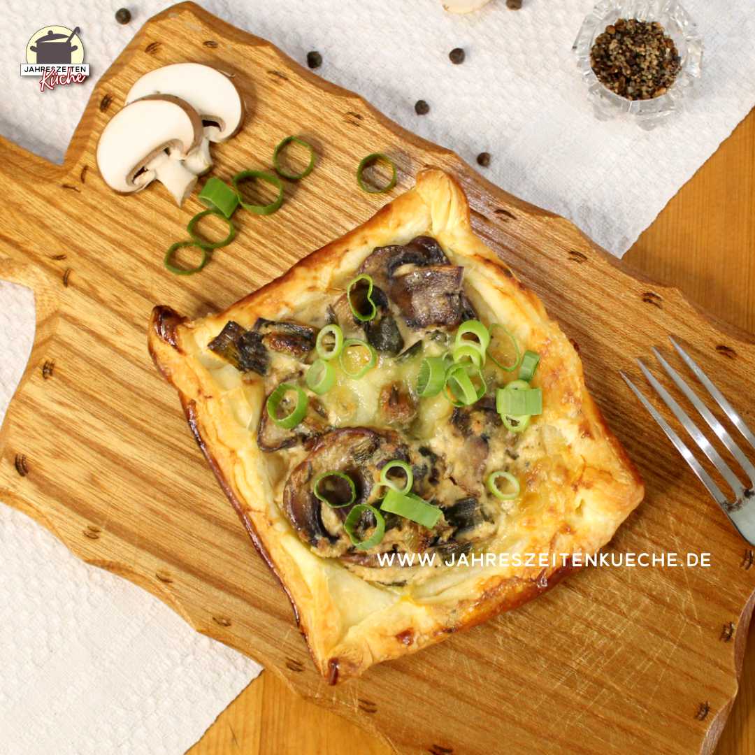 Auf einem Holzbrett liegt ein Champignon-Blätterteig-Snack mit Lauchzwiebeln
