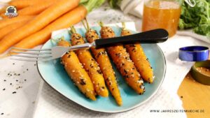 Auf einem türkisfarbenen Teller liegen 5 glasierte Karotten. Quer darüber liegt eine Gabel und im Hintergrund sind frische Möhren