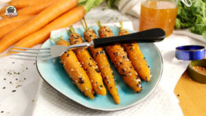 Auf einem türkisfarbenen Teller liegen 5 glasierte Karotten. Quer darüber liegt eine Gabel und im Hintergrund sind frische Möhren