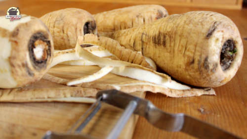 Ein Sparschäler liegt vor ungeschälten und geschälten Pastinaken