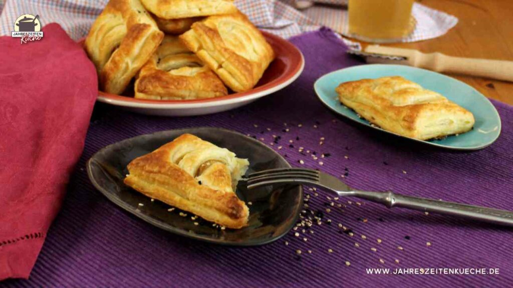 Auf einer lilafarbenen Tischdecke stehen Teller, auf denen Blätterteigtaschen mit Kartoffelfüllung liegen.