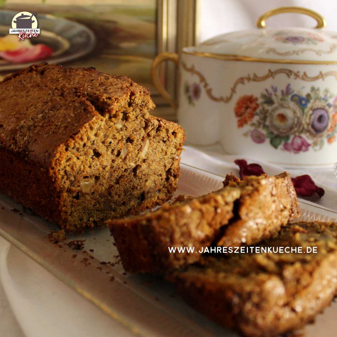 Ein angeschnittener Kastenkuchen mit Karotten und Walnüssen liegt auf einem Glastablett