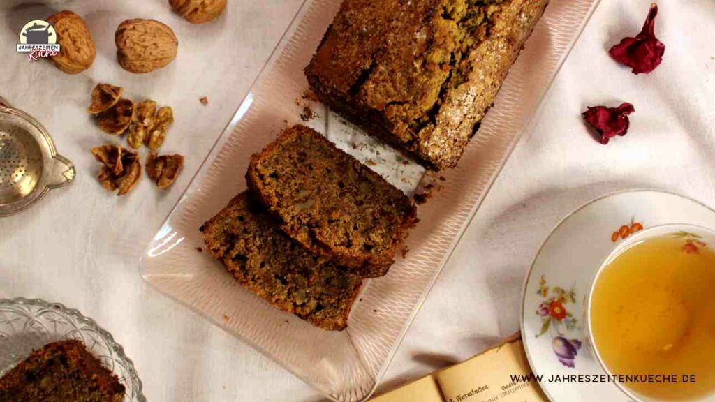 Neben einer Tasse Tee steht ein angeschnittener Karotten-Walnuss-Kuchen auf dem Tisch.