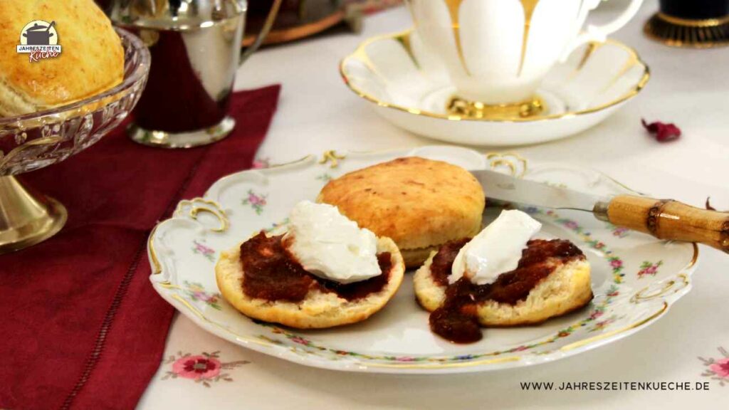 Auf einem zartgeblümten Teller liegen Scones. Zwei Hälften sind mit Marmelade und Clotted Cream bestrichen.