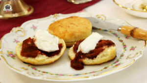 Auf einem zartgeblümten Teller liegen Scones. Zwei Hälften sind mit Marmelade und Clotted Cream bestrichen.