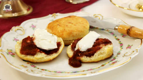 Auf einem zartgeblümten Teller liegen Scones. Zwei Hälften sind mit Marmelade und Clotted Cream bestrichen.
