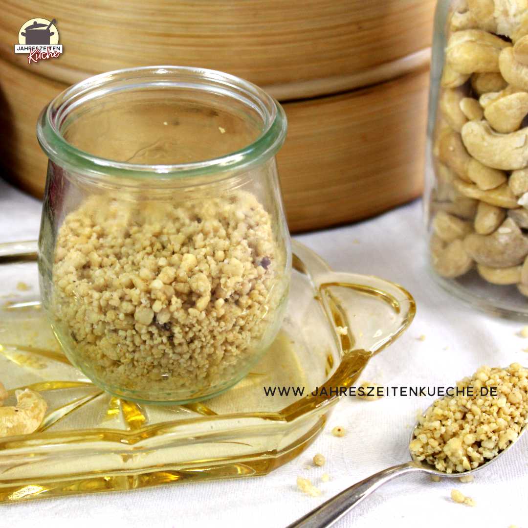 Ein Glas mit veganem Parmesan steht auf einem kleinen Glastablett. Danaben liegt ein Löffel mit veganem Parmesan.