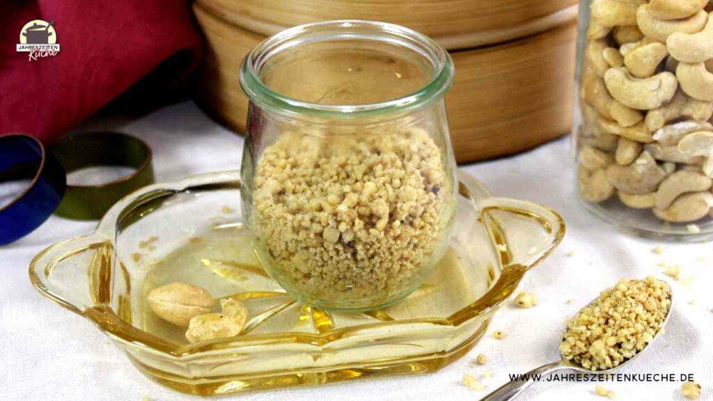 Ein Glas mit veganem Parmesan steht auf einem kleinen Glastablett. Danaben liegt ein Löffel mit veganem Parmesan.