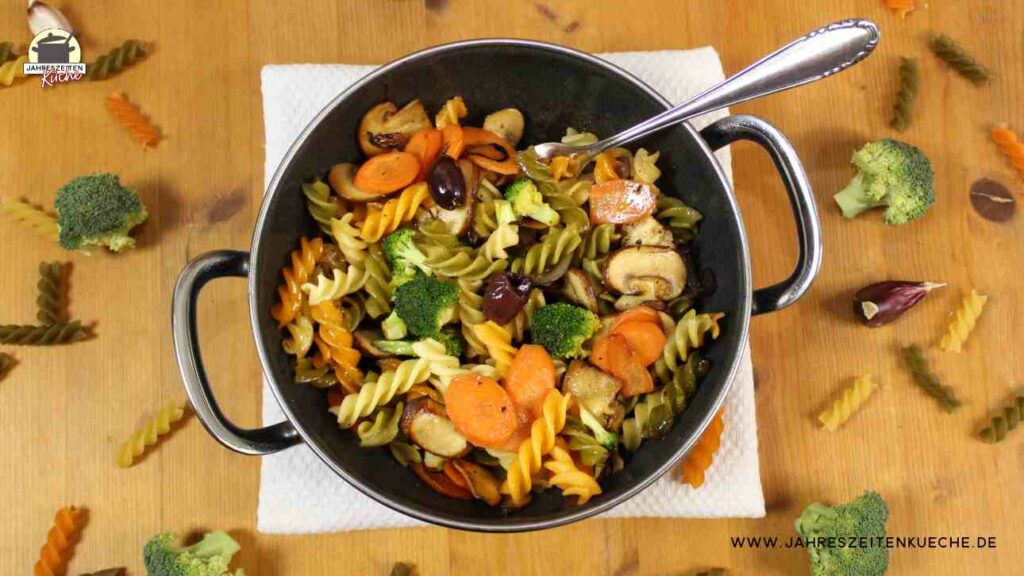 Auf einem Holztisch steht eine Gemüse-Nudelpfanne mit Brokkoli, Champignons und Karotten. Drum herum liegen Spirelli-Nudeln und Brokkoli-Röschen