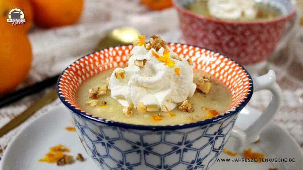 In einer Suppentasse ist eine Portion Petersilienwurzelsuppe mit Orange, Schlagsahne und karamellisierten Walnüssen. Im Hintergrund liegen Orangen