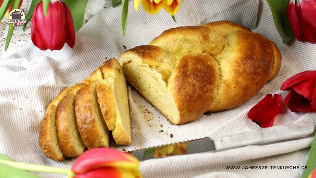 Auf einem Leinentuch liegt ein Hefezopf, von dem vier Scheiben abgeschnitten wurden. Darüber hängen rote und gelbe Tulpen.