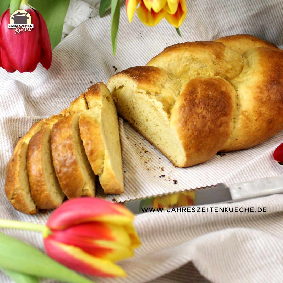 Auf einem Leinentuch liegt ein angeschnittener Hefezopf. Darüber hängen Tulpen.