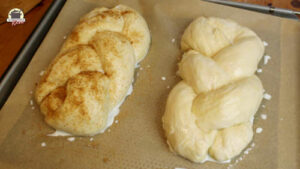 Auf einem Backblech liegen zwei Hefezöpfe. Beide sind mit Milch bestrichen und der andere mit braunem Zucker bestreut.