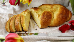 Auf einem Leinentuch liegt ein Hefezopf, von dem vier Scheiben abgeschnitten wurden. Darüber hängen rote und gelbe Tulpen.