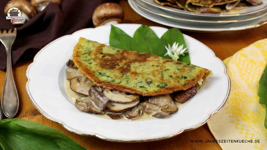 Auf einem weißen Teller liegt ein mit Champignons gefüllter Bärlauch-Pfannkuchen. Im Hintergrund steht ein Teller, auf dem weitere Bärlauch-Pfannkuchen stehen.