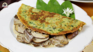 Ein Bärlauch-Pfannkuchen mit Champignons ist auf einem weißen Teller mit frischen Bärlauch-Blättern angerichtet